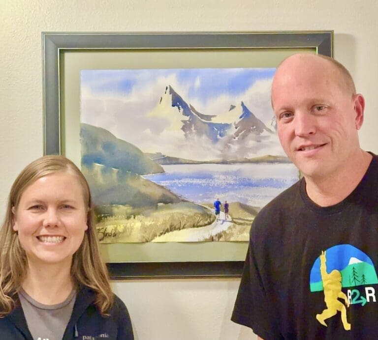 A couple next to their commissioned watercolor painting "Training Together" showing them running long a mountain lake